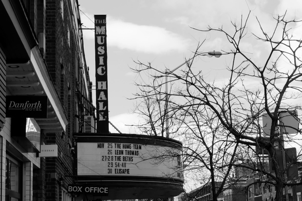 A cold early winter Sunday. Looking up at the marquee of the Danforth Music Hall. Several upcoming gigs are listed. Along the left edge of the frame are the buildings of the street. At right, against the pale, wintry sky are the bare, slender branches of one of the trees planted along the street. 