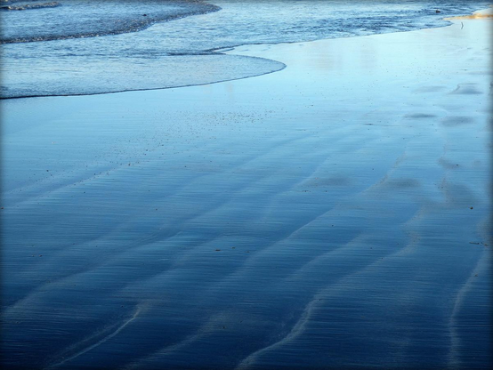 A colour photograph showing the remnants of waves washing ashore over smooth wet sand. The frame is filled with a silken blue hue from the reflection of the blue sky above with detail provides by light ripples of water and sand.