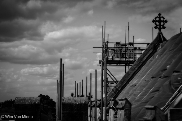Black and white view of the rooftop of the church with an ornate cross.  This is taken during construction work, showing the top section of scaffolding under a fairly dramatic sky full of white-grey clouds.