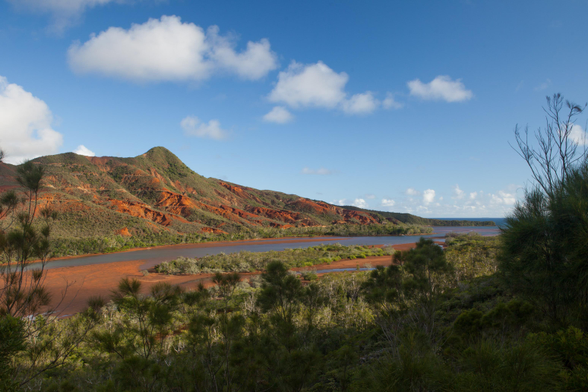 A scenic view of red-hued mountains alongside a river, with lush green vegetation in the foreground and a blue sky dotted with clouds.