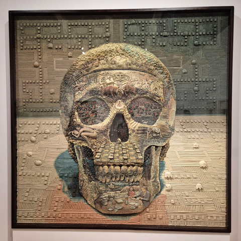 An intricate artwork that looks like a painting but is a carving of a skull that is made of different figures and scenes, mixing reference to pop culture and classical art. 