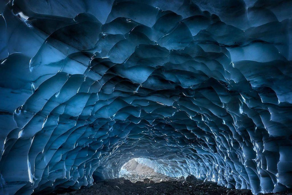 A breathtaking blue ice cave with textured, wavy ice formations leading to a bright opening in the distance. Stone floor visible.