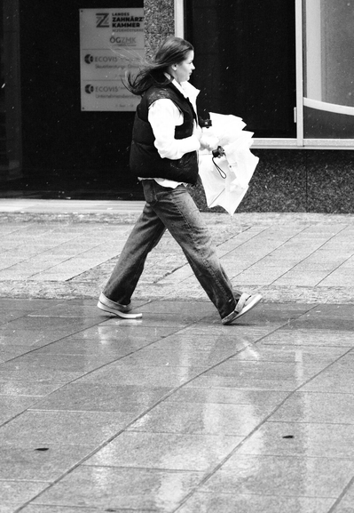 Das Foto zeigt eine Straßenszene in Schwarz-Weiß in der St. Pöltenter Innenstadt an einen regnerischen windigen Tag. Man sieht eine Person mit Regenschirm der durch den Wind verbogen ist und dadurch der Schirm vor ihr ist