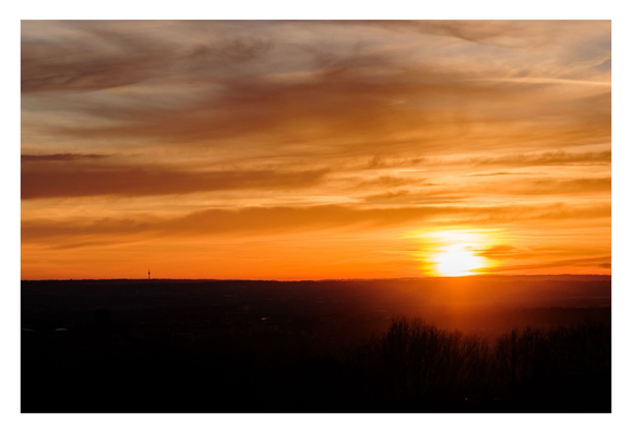 Foto im Querformat.Sonnenuntergang. Der untere Teil des Bildes ist nur schwarze Silhouette der Landschaft. Rechts sind die Kronen einiger Bäume zu erkennen. Am entfernten Horizont geht gerade die Sonne unter. Sie leuchtet gelb. Der Himmel ist knallorange, weiter oben sind viele Wolken. Links am Horizont ist klein ein Fernsehturm zu erkennen.