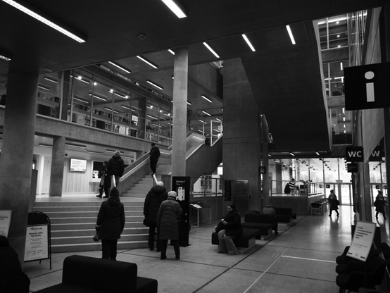 Black and white photograph of modern library lobby interior, with people walking and sitting. Features high ceilings, large windows, stairs, and clear signage.