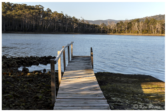A photograph by Kev Peirce showing a small, weathered wooden jetty extending straight out into a calm body of water at Port Arthur, Tasmania.  The jetty is low to the water and surrounded by dark rocks and dark green mossy ground on the shore.  The far bank is lined with dense, tall eucalyptus forest under a clear, pale blue sky, with rolling hills visible in the distance.