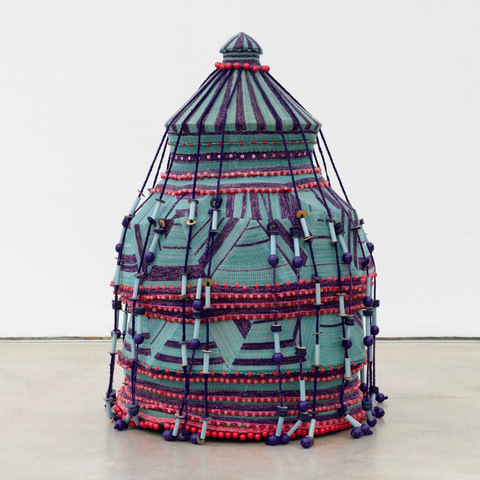 A woven textile sculpture by Jim Drain loosely resembling a large teal and magenta basket with a lid and many long strands or tassels