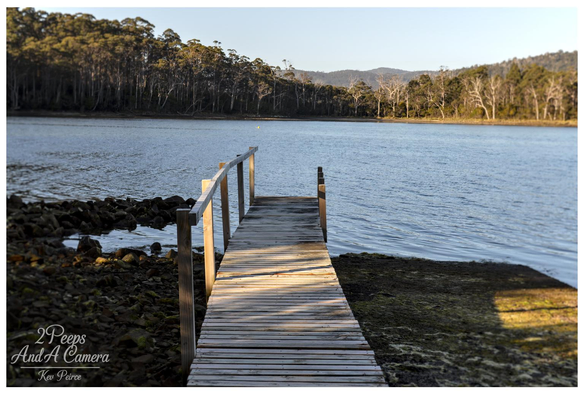 A photograph by Kev Peirce showing a small, weathered wooden jetty extending straight out into a calm body of water at Port Arthur, Tasmania.

The jetty is low to the water and surrounded by dark rocks and dark green mossy ground on the shore.

The far bank is lined with dense, tall eucalyptus forest under a clear, pale blue sky, with rolling hills visible in the distance.