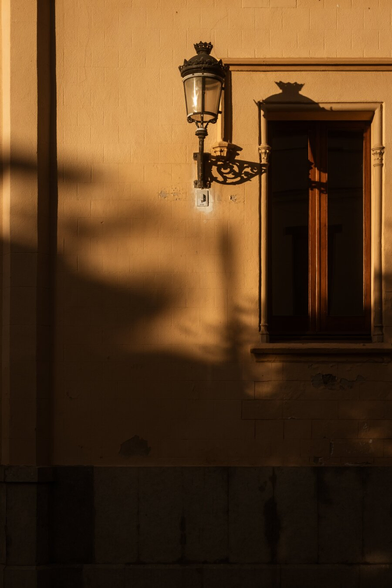 Español: farol clásico de pared proyectando su sombra sobre una fachada color arena, junto a una ventana con marco de madera, iluminado por luz cálida del atardecer.

English: classic wall-mounted lantern casting its shadow on a sand-colored facade, next to a wooden-framed window, lit by warm golden-hour light.