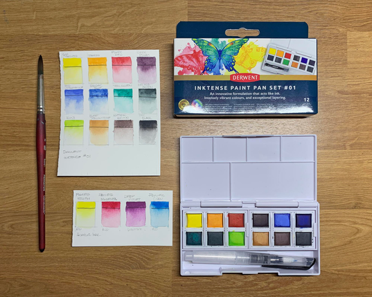 Photo of Derwent InkTense Paint Pan Set #01 along with a small sheet of swatches of the paints, a paint brush, and a second sheet showing swatches of acrylic inks of similar colors