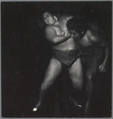 The image is a black and white photograph depicting two sumo wrestlers engaged in what appears to be an intimate moment, possibly after or during their training. The wrestler on the left has his eyes closed with a relaxed expression while leaning back into the embrace of the other wrestler. He wears only a traditional mawashi (sumo belt), revealing his upper body and part of his legs. His physique is robust, typical for sumo wrestlers known for carrying excess weight around their midsection.

The second wrestler has an intense focus in his gaze as he leans forward with one arm wrapped tenderly around the first wrestler's waist or back. This individual also sports a mawashi but wears dark shorts over it, and they are partially obscured by shadows, which adds to the dramatic effect of the image.

Both wrestlers' skin tones contrast against the monochromatic background, drawing attention to their physical presence. The lighting highlights certain areas on their bodies, creating soft contrasts that suggest movement or tension in an otherwise still scene. There is no visible text overlaying the photo, and it captures a candid moment with raw human emotion conveyed through body language.

The photograph seems to be from Ed van der Elsken's collection as referenced by the provided link, which suggests this image may have been taken during one of his travels or assignments in Jap [...]
