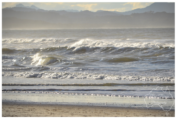 A sweeping, elevated view of a sandy beach on the Northern Rivers coast of New South Wales (NSW), south of Byron Bay.   Powerful white waves roll in from the deep blue ocean, meeting the golden sand. Coastal scrub and a wooden fence line the foreground, with a distant rock breakwater visible.