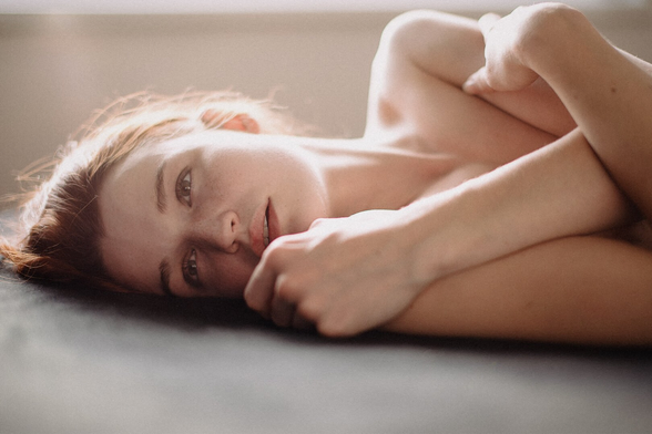 Soft, natural-light portrait of a woman lying on her side, with gentle shadows and a calm, intimate expression against a neutral background. The woman has red hair and freckles.