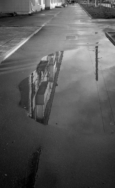 Black and white photo. A close-up shot of the road: wet asphalt, a building reflected in a puddle in the middle.