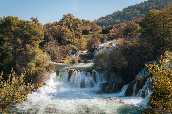 A wide waterfall flows into a river surrounded by forest. Trees fill the background, and mist floats in the air.