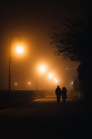 Two people holding hands, walking down a foggy street with streetlamps.