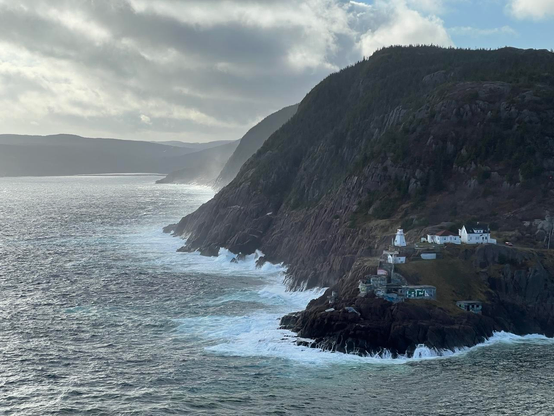 A rugged steep coastline provides a backdrop for lighthouse perched on a low bump of land surrounded by mist from breaking waves. The cloud filled sky is grey and backlit by the sun, with one small patch of blue in the upper right corner. The barren rocky coast itself has muted colours in the distance due to the breaking waves, giving the effect of a glow. The lighthouse is surrounded by other low structures, including a graffiti covered bunker below and a white house and outbuildings to the right. [St. John’s (NL, Canada), November 2025]