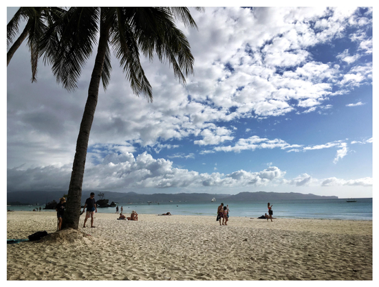 Photo of a wide island beach under a blue sky half-covered with cumulus clouds. At left is a tall, backlit palm tree, its fronded leaves pushing past top frame. The sand is textured with multiple little dunes, ripples and indentations. Tiny figures of people stroll along the beach. Beyond is a strip of blue sea, with a line of low mountains on the horizon.