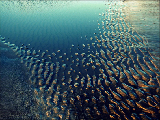 A colour photograph showing standing water over rippled sand in late afternoon winter sun with a pool of water reflecting the blue sky above. Rippled sand passes diagonally through the frame from bottom right until it reaches a deep turquoise pool of standing water at the top of the frame.