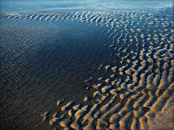 A colour photograph showing an arc of rippled sand at low tide bordering a pool of standing water. The sand arcs through the right of the frame towards the top of the photograph partially surrounding the standing water at the left of the frame.