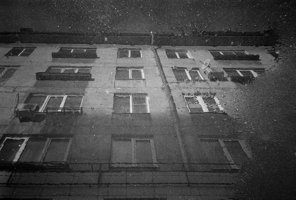 In the black-and-white photo, a multi-storey building is reflected in a puddle.
