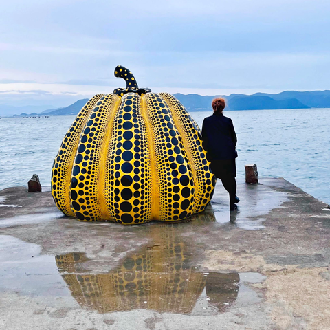 Ein Kunstwerk am Meer: Ein großer gelber Kürbis mit vielen schwarzen Punkten unterschiedlicher Größe, auf einem nassen Anleger, der ins Meer führt. 
An der rechten Seite steht eine rothaarige Frau, kleiner als der Kürbis. Sie ist schwarz gekleidet, und lehnt sich zum Meer gewandt an den Kürbis.