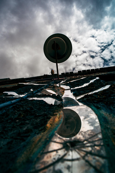 The image presents a low-angle perspective of an outdoor scene, dominated by a street lamp and the sky. The composition is dramatic, utilizing a strong vertical perspective to emphasize the height of the lamp and the vastness of the sky.

The lamp is mounted on a wall with a rough, textured finish. The surface is primarily dark in color, with areas covered in broken mirror pieces, creating a distorted reflection of the lamp. 