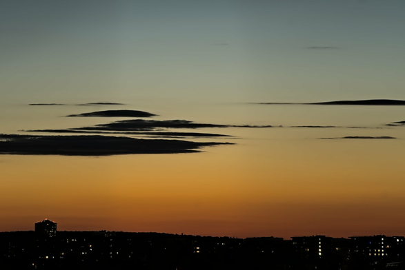 Im Vordergrund sind die dunklen Umrisse von Gebäuden zu sehen, die sich deutlich vor einem farbenfrohen Himmel abheben. Der Himmel ist in warmen Orange-, Gelb- und Blautönen des Sonnenuntergangs gehalten. Über dem Horizont erkennt man sanfte Wolkenformationen, die vom Licht der Sonne angestrahlt werden. Die Silhouetten der Gebäude und Wolken sind dunkel und bilden einen starken Kontrast zum leuchtenden Himmel. Das Bild wirkt sehr stimmungsvoll und ruhig.