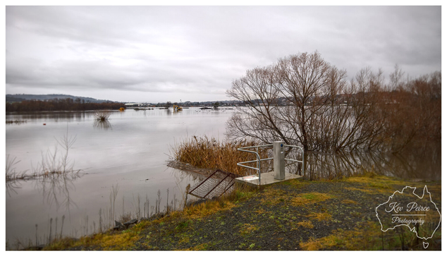 A wide angle landscape photo on a grey, overcast day showing significant flooding over the riverbanks.  The foreground features a small metal railing fixture on a patch of grassy dirt, leading down to the wide, muddy floodwaters.  Submerged bushes and trees are visible across the water, with construction equipment visible further in the distance.