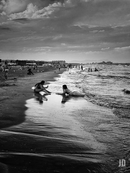 Fotografía en blanco y negro de una playa concurrida bajo un cielo nublado y dramático.
Se observa la orilla mojada con reflejos nítidos de las figuras humanas. Dos mujeres jóvenes son el foco central: una está sentada y la otra tumbada en la arena, justo donde rompen las pequeñas olas.
La línea de costa se extiende hacia el fondo, donde se distinguen edificios, estructuras de playa y montañas lejanas. El mar presenta un oleaje suave.
El uso del blanco y negro resalta las texturas de la arena mojada y las formaciones de las nubes, creando una sensación de atardecer o día tormentoso.
