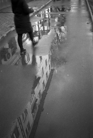 In the black-and-white photo, a multi-storey building and a walking man are reflected in a puddle.