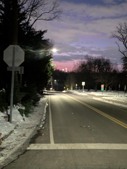 Photo looking down a road toward the east and Center City, Philadelphia. The early morning blue sky is streaked with clouds. It glows light pink and red just above the horizon. The lights of downtown Philadelphia's tallest buildings also appear above the horizon. There are street signs on both sides of the road, a solid double yellow line down the center and snow on either side.