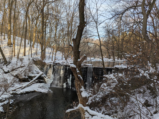 Photo of a waterfall with ice along the edges and on the water above the drop and snow covering the stream banks. The trees are bare and the sky is light blue.