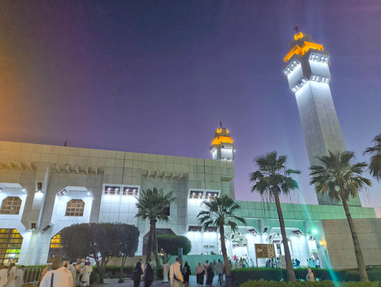 A wide-angle photograph taken at twilight shows the exterior of a large, modern mosque. The building has white stone walls and windows glowing with warm light. Two tall minarets with distinctive geometric tops and warm lighting stand against a deep blue and purple sky. In the foreground, numerous pilgrims dressed in white Ihram clothing walk along paved pathways amidst palm trees and manicured hedges.