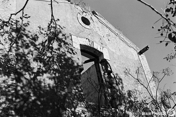 A monochrome photograph of a weathered building with a partially collapsed wall and a large window frame, surrounded by overgrown branches and shadows, showcasing an abandoned and decaying structure against a clear sky.