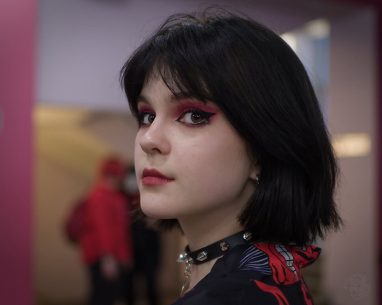 The image captures a young woman in profile, her gaze directed off to the side. She has short black hair and is wearing bold makeup with red eyeshadow that matches her vibrant lipstick. Her outfit includes a choker adorned with silver studs around her neck and she's dressed in a blouse featuring an intricate design on one shoulder.

The background suggests an indoor setting, possibly a public space like a museum or gallery, indicated by the blurred figures of people moving about behind her. The overall mood is contemplative and artistic, enhanced by the contrast between the clarity with which the woman is portrayed against the soft focus of the backdrop.