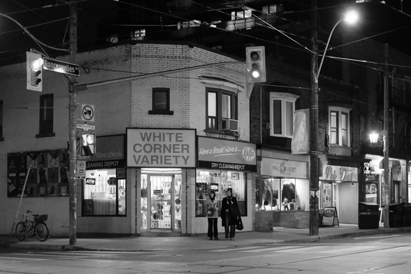 Night. Wide shot of a small convenience store on the diagonal corner from the camera. The sign reads “White Corner Variety”. At far left, a bike is parked beneath a mural on the building’s side. At right, the lights of the buildings along Roncesvalles Avenue lead into the distance. Outside the store, two warmly dressed pedestrians wait for the light to change.
