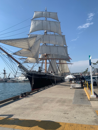 A tall ship with white sails is docked at a harbor under a clear blue sky. The ship's black hull contrasts with the surrounding waterfront, where a small market stall is visible on the pier. Additional boats can be seen in the background.