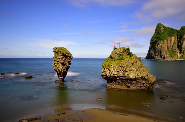 Photo couleur d'un paysage marin où l'on voit au premier plan une petite langue de sable puis la mer où l'on voit deux rochers qui sont considérés comme des divinités dans la religion shinto (ils sont nommés d'après leur ressemblance avec deux dieux de la religion shinto.Le plus petit des rochers à une forme particulière. La base est érodée et forme une pointe qui s'évase un peu jusqu'à son sommet qui n'est pas très haut. Le 2ème rocher a accroché à son sommet un peit tori rouge (portail shinto). Plus loin à droite,  de la photo, une grande falaise. Le ciel est bleu avec quelques nuages.