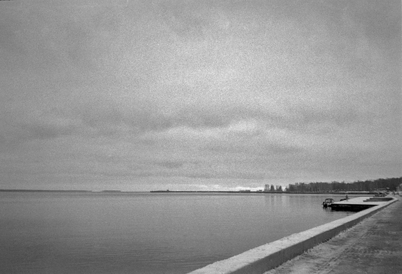 The black-and-white image shows part of the embankment shore and the water surface of Lake Onega. The sky is covered with clouds.