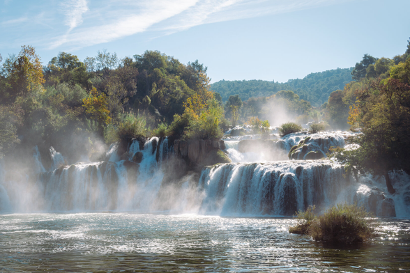 A wide waterfall flows into a river surrounded by forest. Trees fill the background, and mist floats in the air.