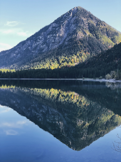 Ein Berg mit Spiegelbild im blauen Wasser eines Sees und mit einem grünen Streifen sonnenbeschienener Bäume