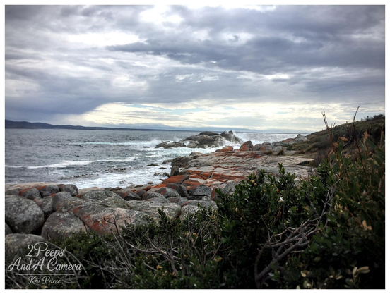 Dramatic landscape photo of Binalong Bay, Tasmania, featuring a wide expanse of granite boulders covered in vibrant orange lichen extending into the sea.

Waves are crashing over the outer rocks under a dark, heavy, and cloudy sky. Green coastal scrub dominates the foreground on the right.
