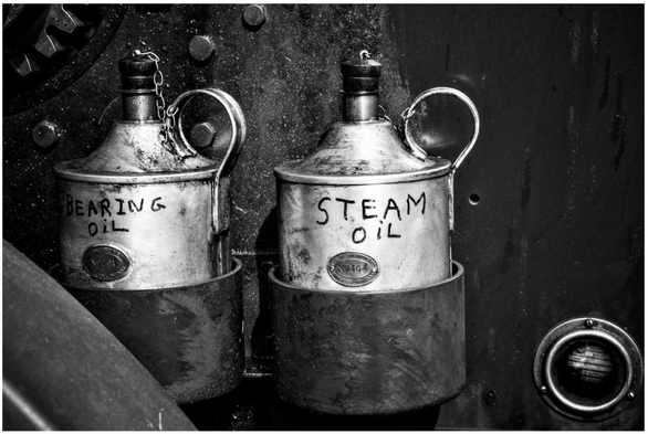 A black and white, close-up photograph of two weathered metal oil cans, or lubricators, mounted side-by-side on a dark, textured metal surface. The can on the left is hand-labeled with the words 'BEARING OIL' and the one on the right is labeled 'STEAM OIL'. The image focuses on the worn, grimy details of the canisters and their practical, utilitarian purpose on the vintage machinery.