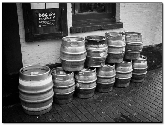 A stack of metal beer kegs arranged against a wall outside a pub. The kegs vary in size and are positioned in a slightly irregular manner, creating a visually interesting composition. In the background, there is a window with a sign indicating that the establishment is dog-friendly. The image is presented in black and white, highlighting the textures of the kegs and the cobblestone pavement.