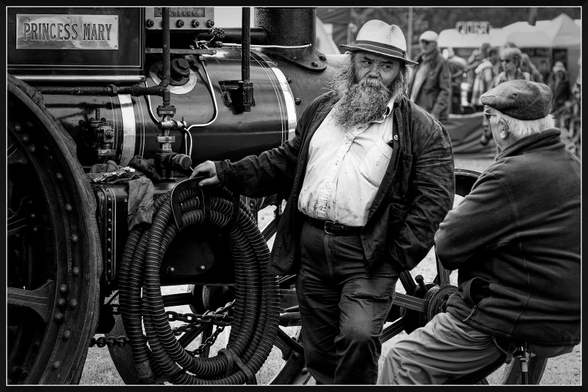 Monochrome image of two older men sharing an easy conversation beside a traction engine named Princess Mary at a steam fayre. One, with a full grey beard and straw hat, leans against the machine with a relaxed confidence, while the other, in a flat cap and jacket, sits perched on the wheel. Their presence and camaraderie give the scene a timeless, almost nostalgic quality, as if guardians of both memory and machine.