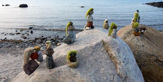 Kleine Steinfiguren mit Tang als Haare stehen auf zwei Felsen. Im Hintergrund Meer.