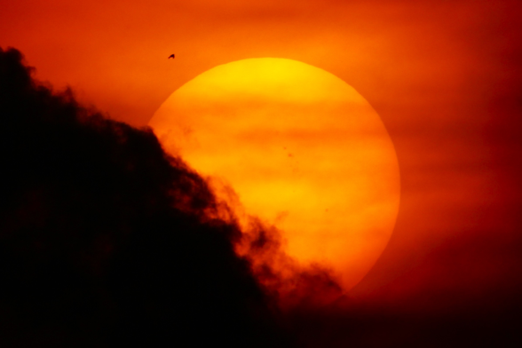 Sunset photograph of a big yellow and orange sun in a dark orange sky with a dark cloud billowing up from the lower left.
