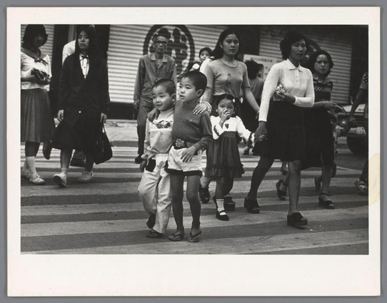 The image captures a moment of pedestrians crossing an urban street at what appears to be a pedestrian crosswalk. The scene is in black and white, suggesting it may have been taken some years ago or intended for artistic effect. Multiple figures are visible: adults and children alike. Two young boys appear to be the focus; they stand close together with one boy seemingly leading while holding onto the other's arm, possibly indicating a sibling relationship or guidance from an older child towards safety across the road.

The adult women accompanying them wear different styles of clothing that suggest varied roles within their community—possibly mothers and caregivers. They are dressed in skirts, blouses, and cardigans which indicates this photo might have been taken during cooler weather periods like spring or fall.

There's a sense of everyday life captured here as people go about their day walking across the street with no immediate urgency, perhaps after school hours given that some children wear what seem to be uniforms. The expressions on their faces are difficult to discern due to the resolution and black-and-white rendering but generally exude normalcy in daily activities.

The background is not very detailed; however, a building with large windows or possibly storefronts can be seen behind them suggesting this photo was taken downtown or near commercial establishme [...]