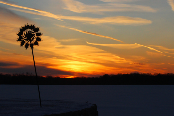 Sunset photograph of yellow and orange clouds over a frozen lake. A wind-spinner stands in silhouette to the left with a shape like a round flower.